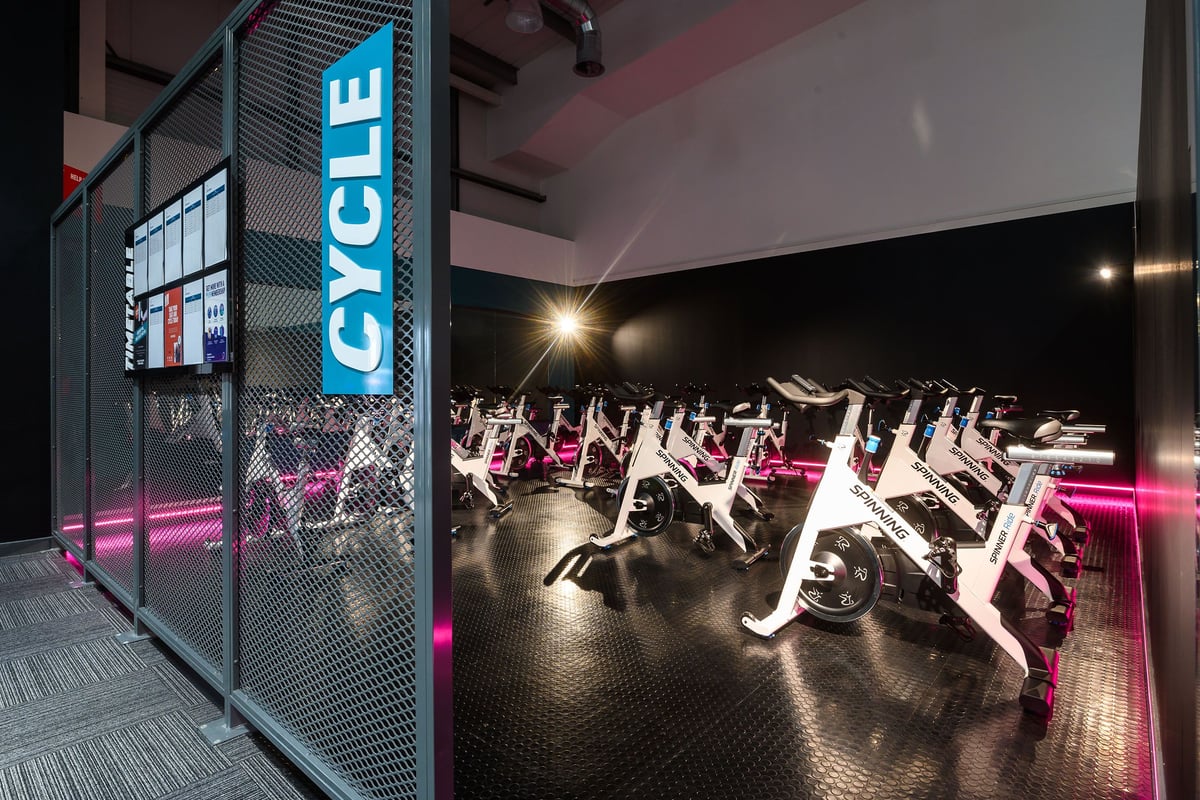 PureGym Bristol Abbey Wood Retail Park: Row of stationary cycles, mirrored wall, vibrant neon lighting.