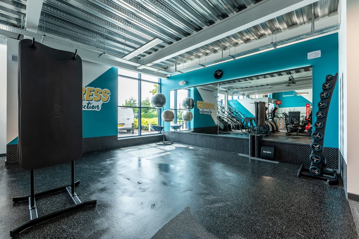 PureGym Coleraine: Gym studio with freeweights, speakers, rubberized flooring, and mirrored walls in teal decor.