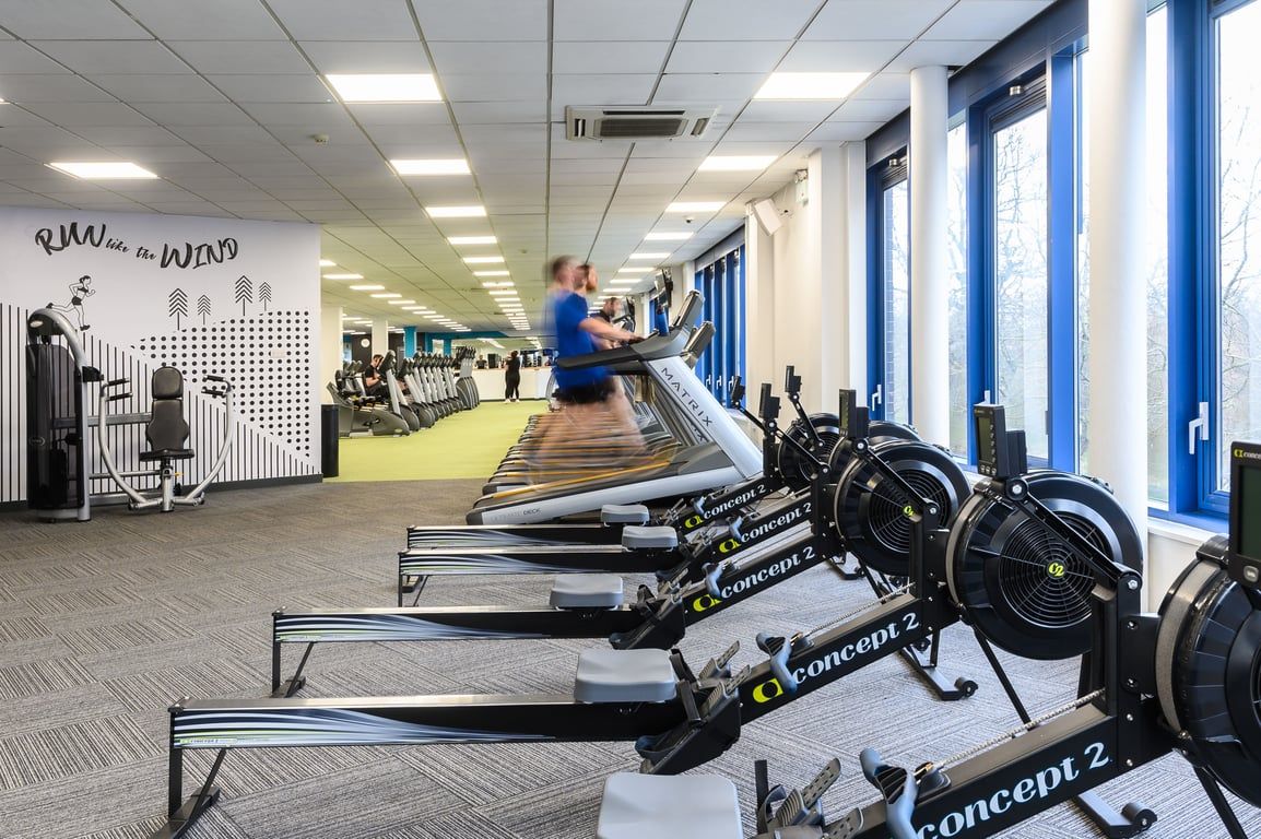 PureGym Edinburgh West: Rows of stationary bikes, treadmills, weight benches with dumbbells in a gym corridor.
