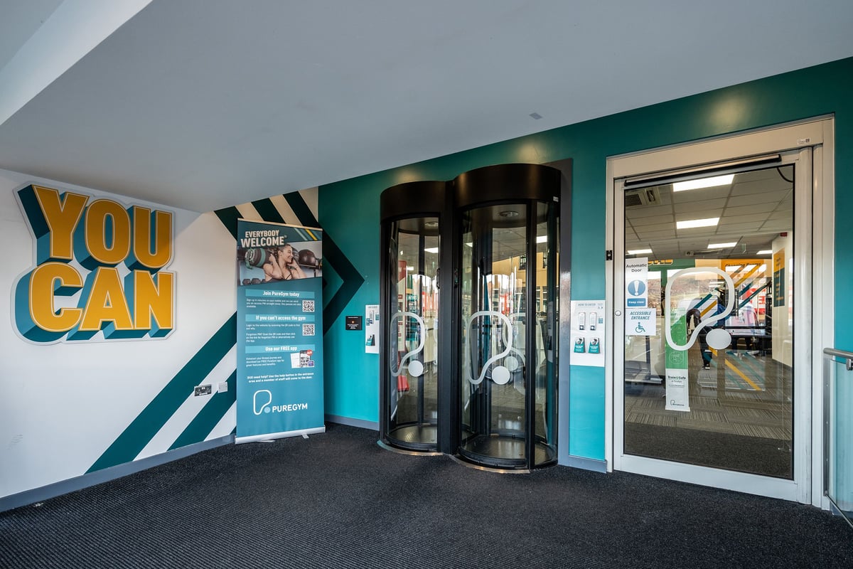 PureGym Belfast Boucher Road: Glass revolving doors, "You Can" signage, fitness advertisement posters.