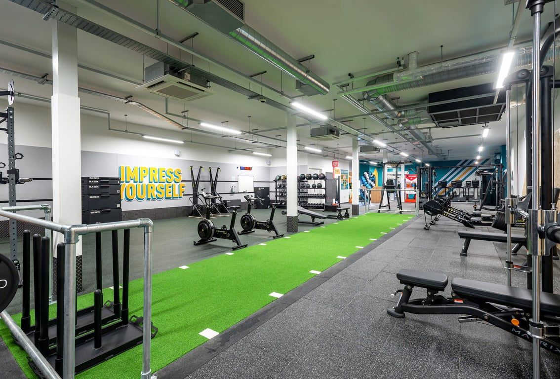 PureGym Stratford-upon-avon: Weight racks, benches, green turf area, strength training equipment, and motivational signage in a gym setting.
