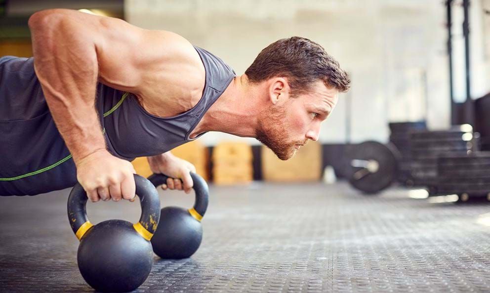 Man doing push-up using kettlebells in a gym with weights in the background
