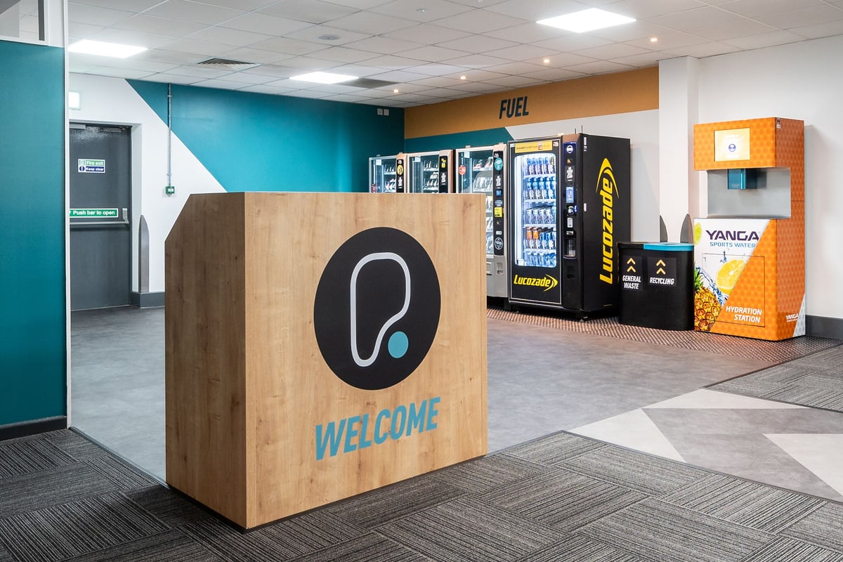 PureGym Harlow: Vending machines, welcome sign, teal and orange colour scheme in a lobby area.