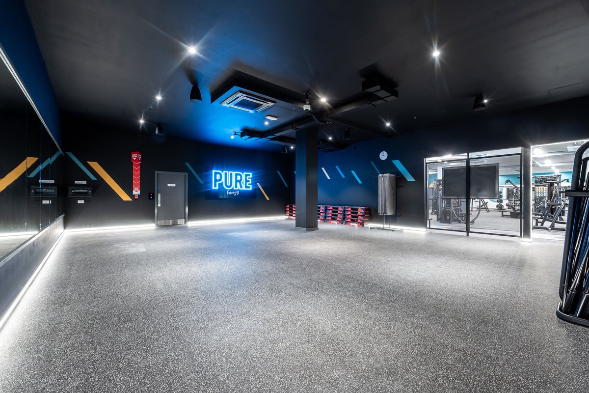PureGym Coalville: Neon-lit studio with rubberized flooring, sleek equipment, and mirrored walls.