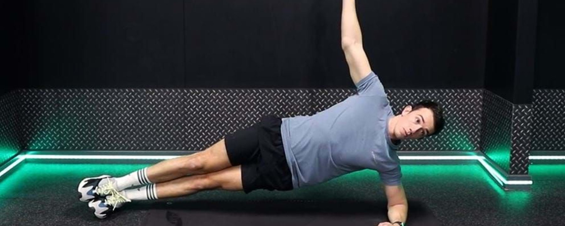 Man in sportswear doing side plank exercise on a mat in a gym room with green lights