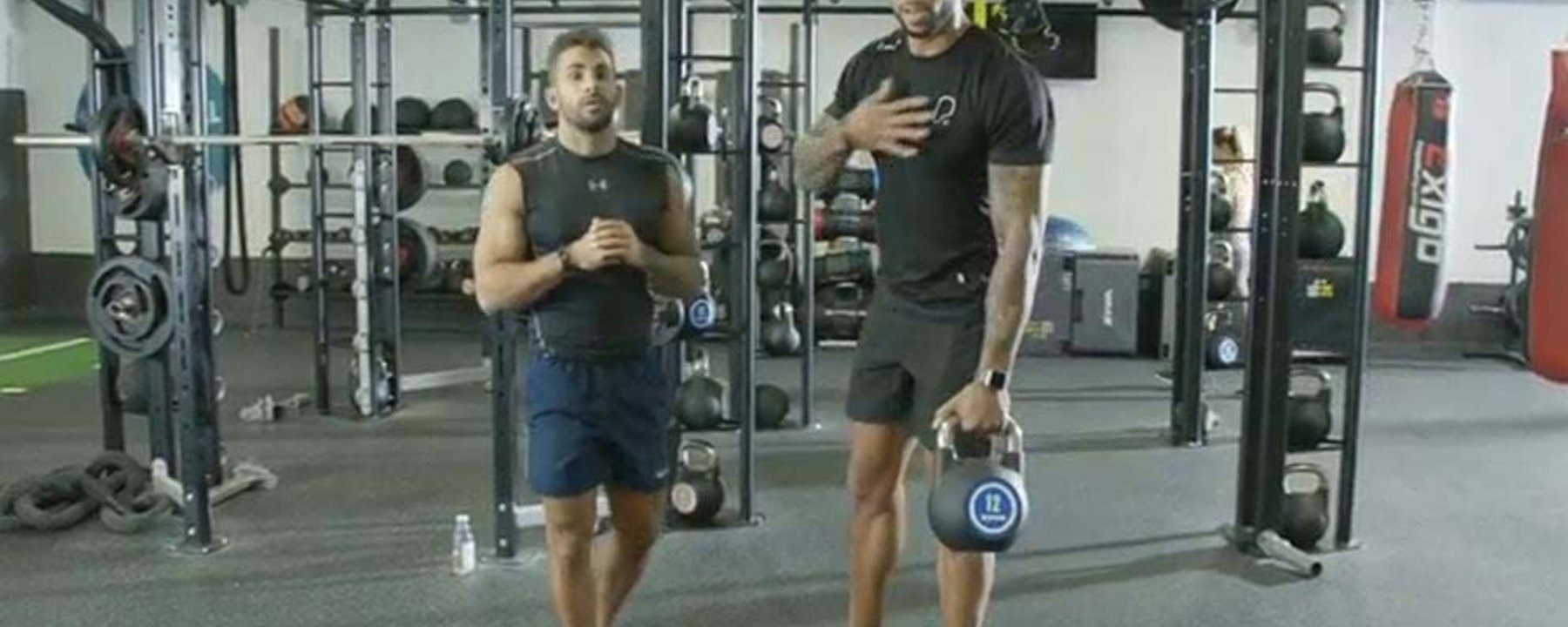 Two men in gym gear standing in a gym with kettlebells and weights around them