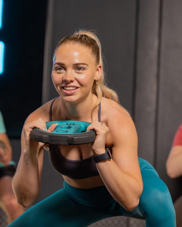 Energische Frau lächelt, während sie eine Gewichtsscheibe in einem modernen Gym-Studio mit blauer Beleuchtung hält