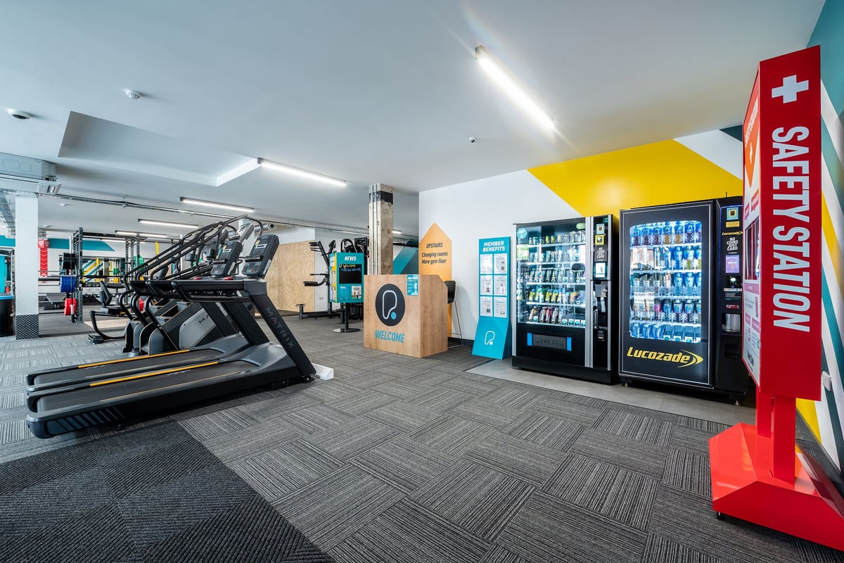 PureGym London Welling: Treadmills, exercise bikes, vending machines, stairs, and safety sign in a modern fitness facility.