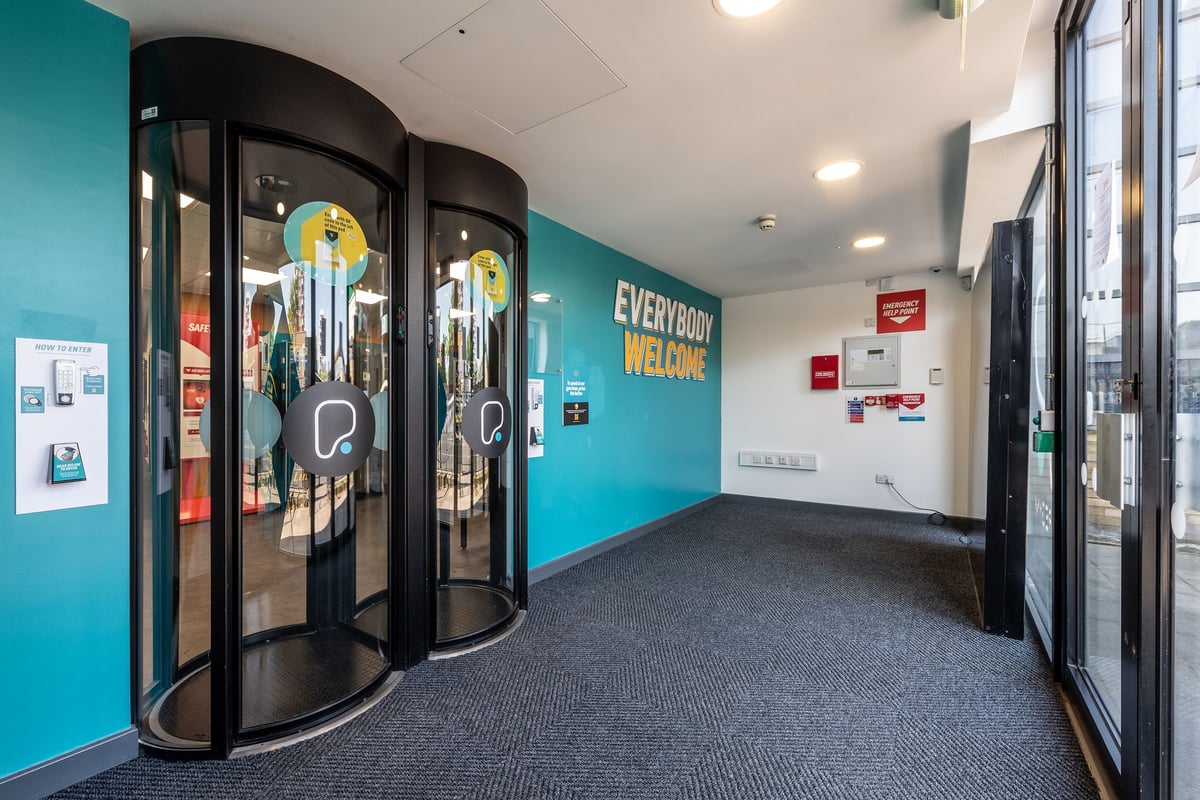 PureGym London Limehouse: Glass revolving doors, blue-teal walls, "Everybody Welcome" signage - entry area of a gym.