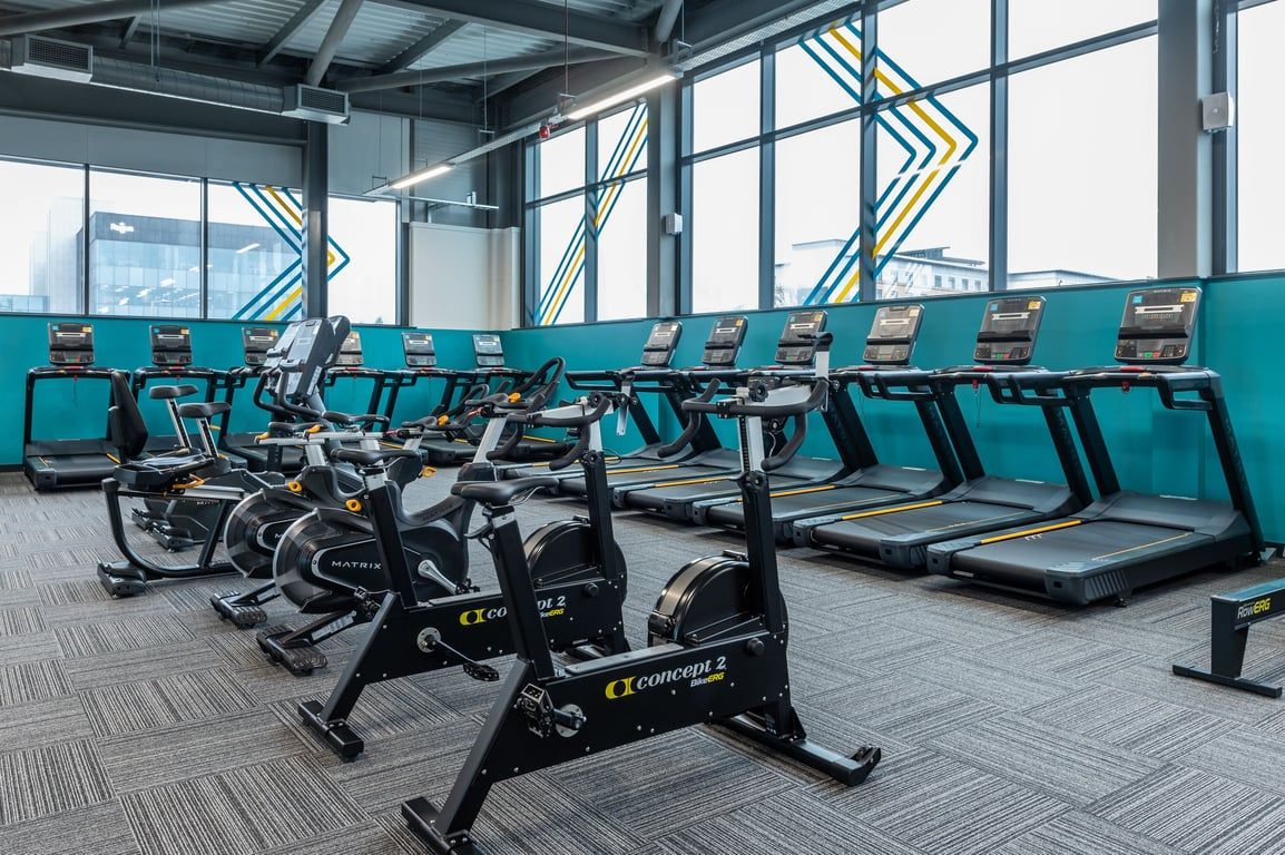 PureGym Plymouth Derriford: Rows of stationary bikes and treadmills in a bright gym space with large windows.