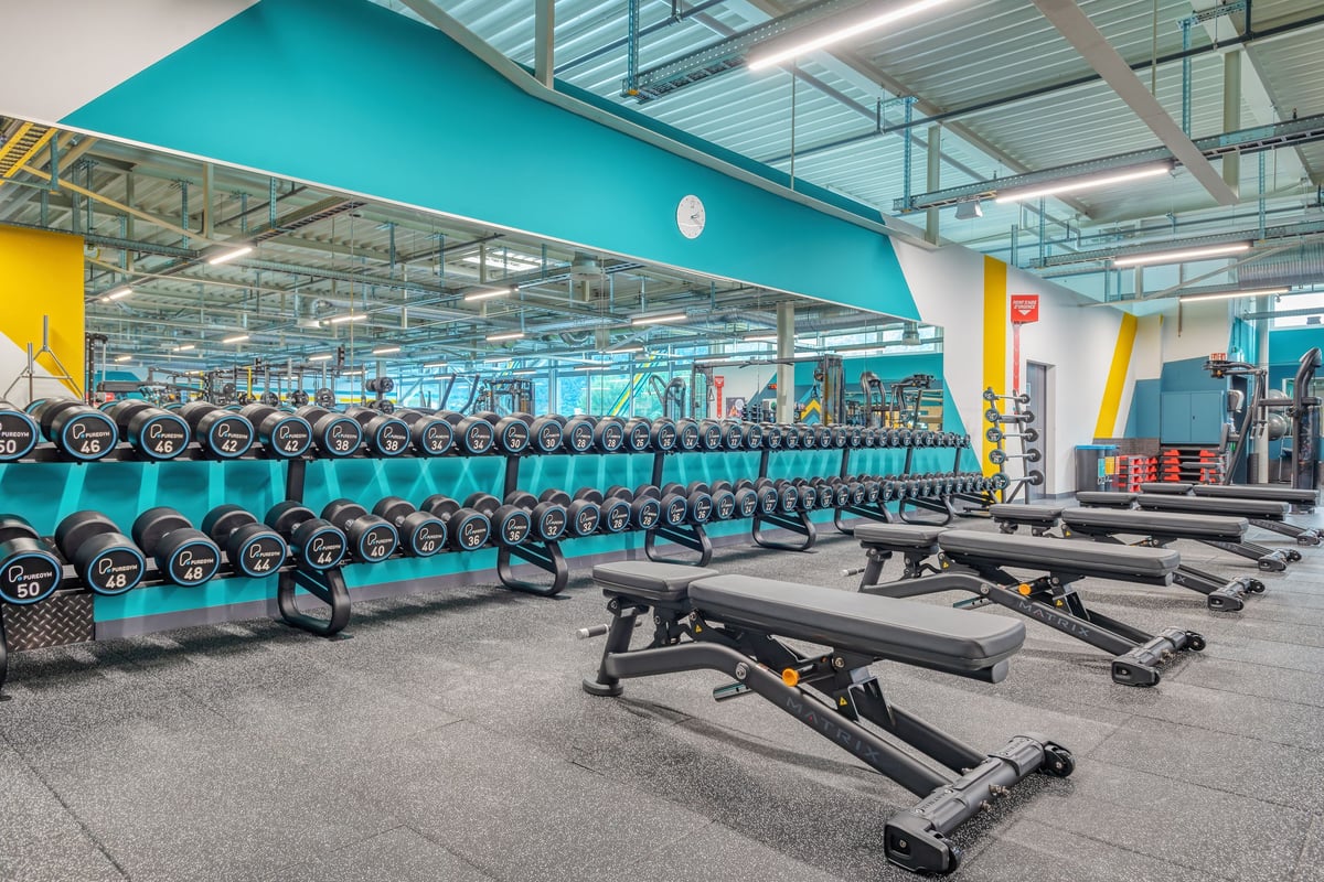 PureGym Martigny: Reihen von Hantelbänken, Hanteln in Regalen und gummierter Bodenbelag.