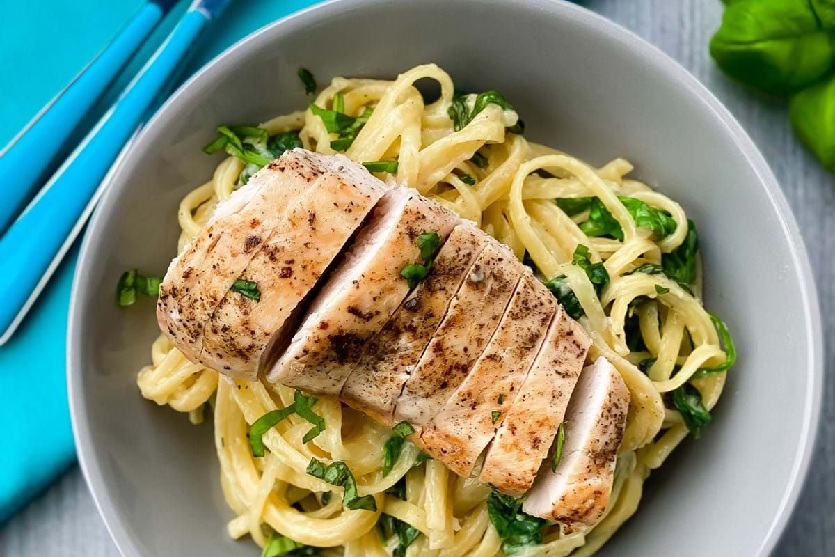 Grilled chicken breast slices on creamy pasta with spinach in a grey bowl with cutlery.