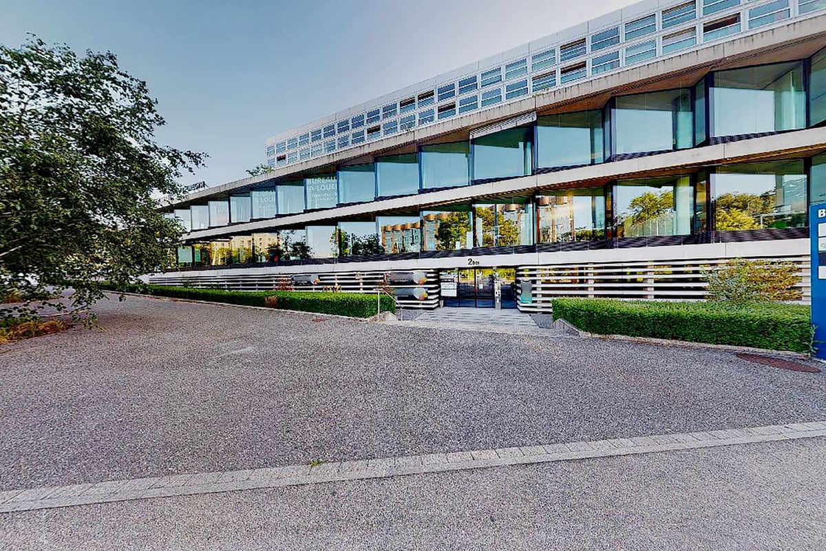 PureGym Genf Le Bouchet: Modern office building with large windows, landscaped grounds and parking area in front.