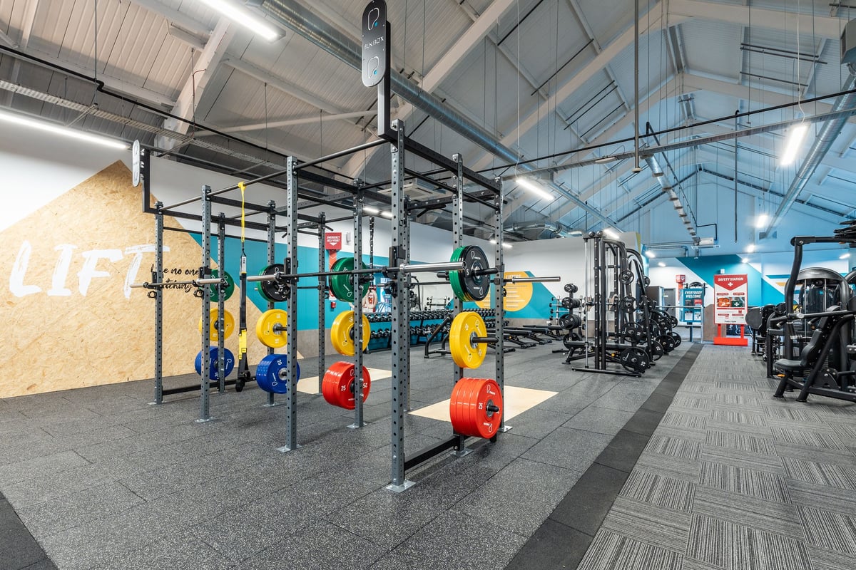 PureGym Kendal: Squat racks, weight plates of various colours, rubberized flooring in a bright gym studio.