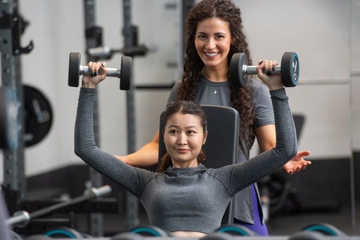 Woman lifting dumbbells on a bench press with a trainer assisting her in a gym