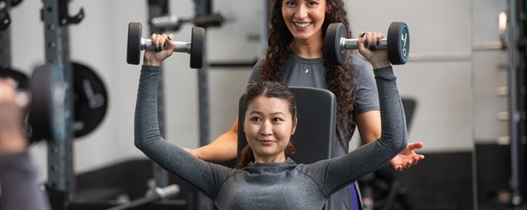 Woman lifting dumbbells on a bench press with a trainer assisting her in a gym