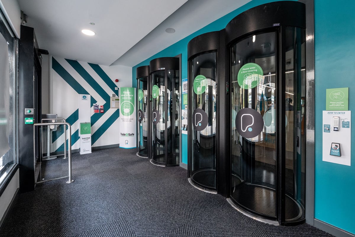 PureGym London Sydenham: Glass-encased revolving entry portals with signage in a turquoise fitness studio environment.
