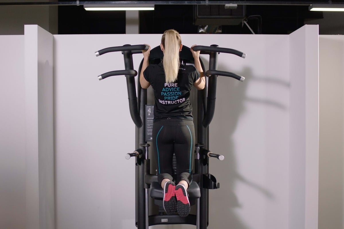 Woman doing pull-ups on a gym machine wearing black workout clothes and pink-soled shoes