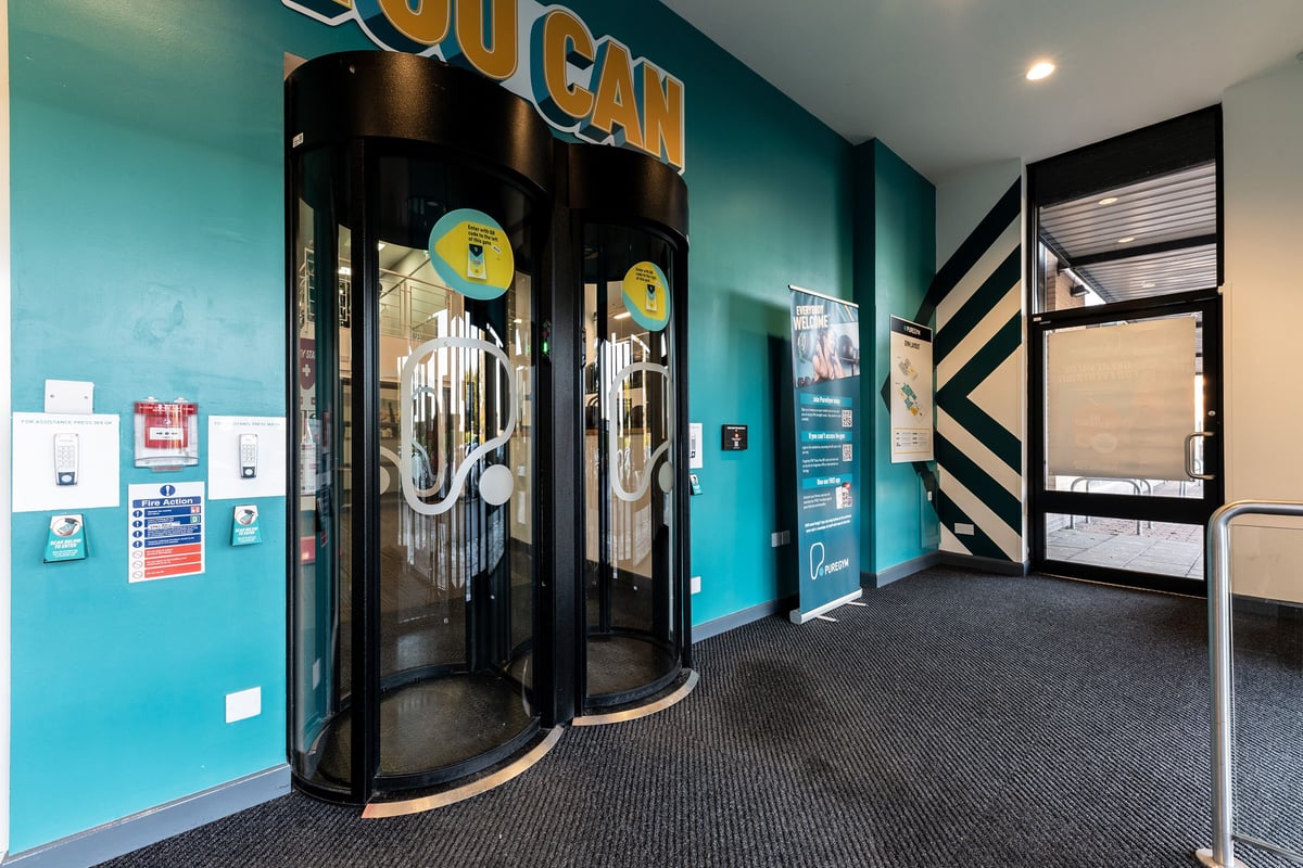 PureGym Bangor Northern Ireland: Revolving doors, vibrant teal walls, signage reading "Gym" with exercise equipment icons.