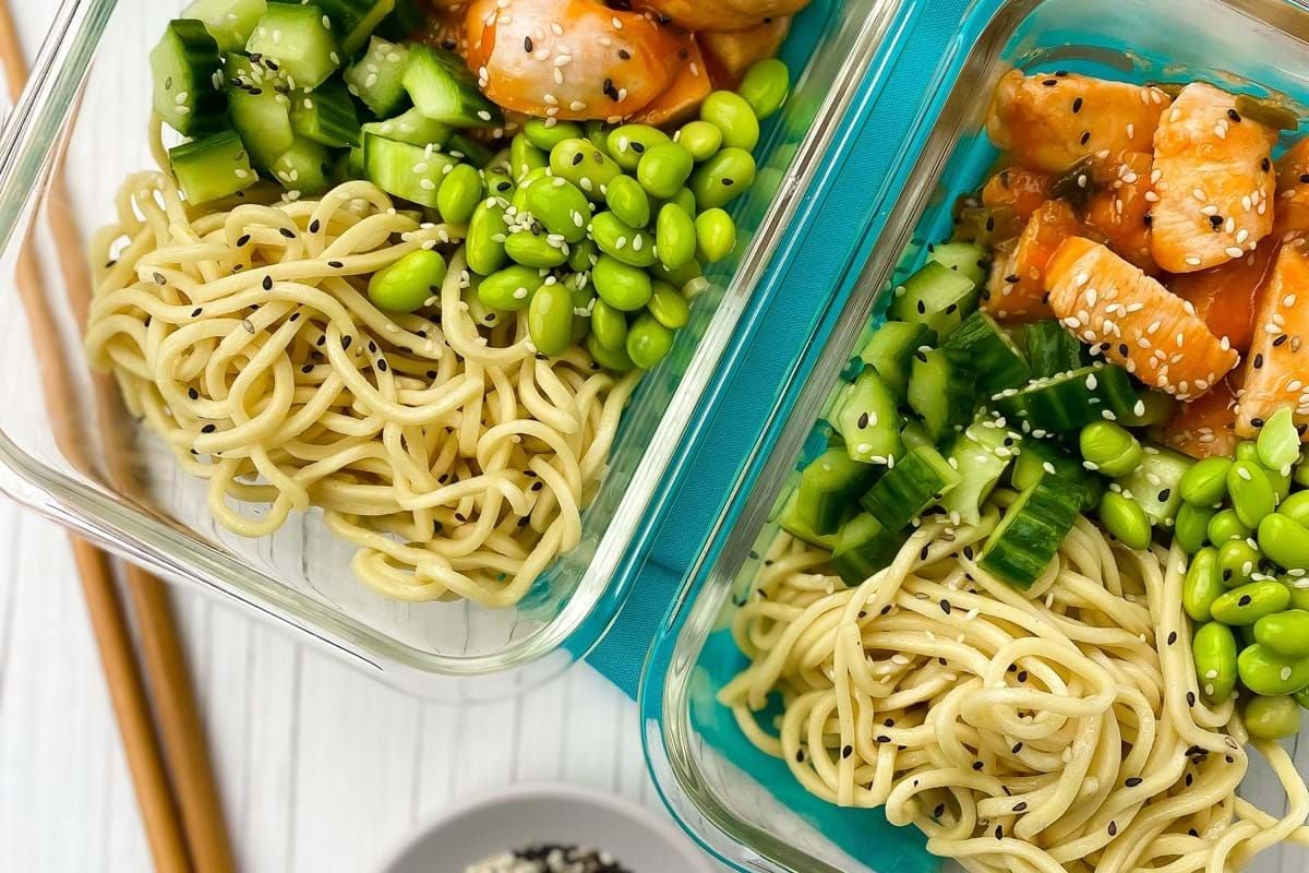 Two glass containers with noodles, diced cucumber, edamame, and sesame chicken pieces