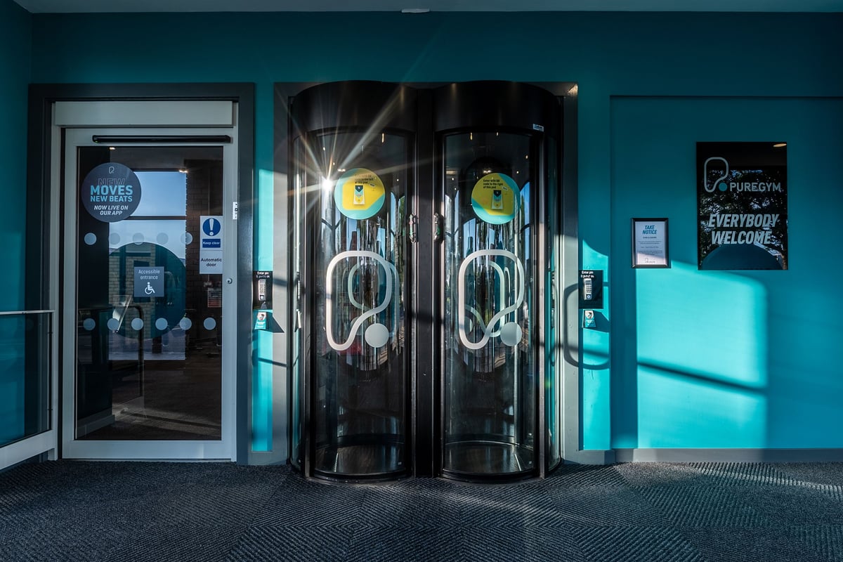 PureGym Weston-super-mare: Two tall cylindrical automated entry turnstiles in a teal-colored gym lobby.
