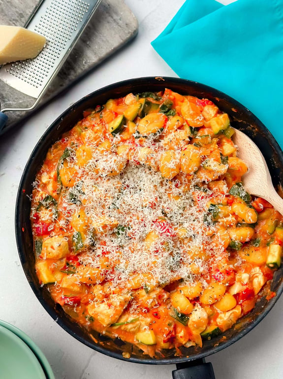 Pan of gnocchi with vegetables and grated cheese on top next to cheese grater and blue nap