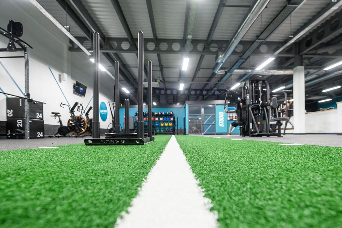 PureGym Leeds Thorpe Park: Indoor gym setup with turf area, weight racks, functional training rigs, and large display screen.