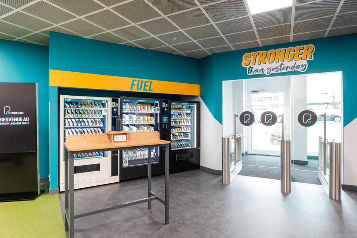 PureGym Lausanne Prilly: Snack and drink vending machines, turnstiles at entrance, bright teal and yellow facility interior.