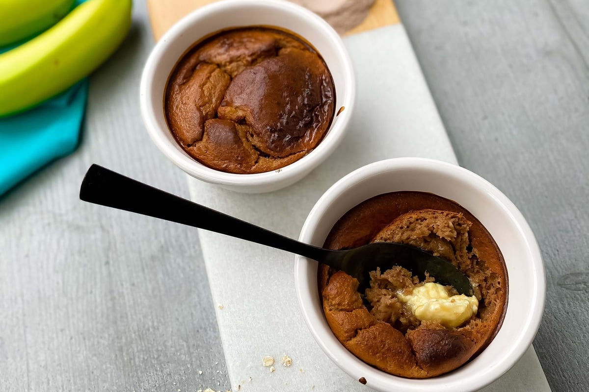 Two baked oat puddings in white ramekins on a marble board with bananas and oats nearby