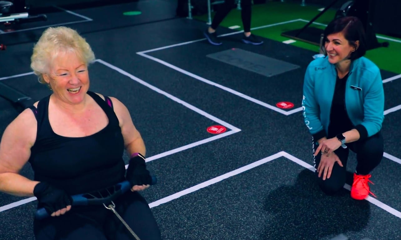 Older woman using rowing machine while younger woman kneels beside her smiling in gym
