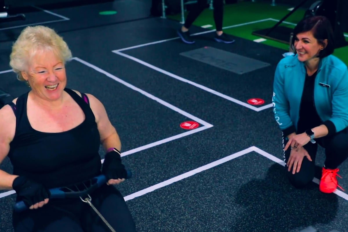 Older woman using rowing machine while younger woman kneels beside her smiling in gym