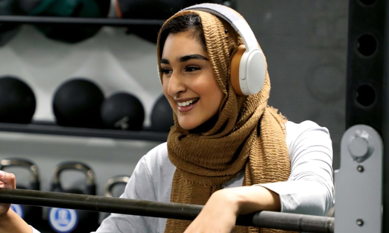 Smiling woman wearing a brown hijab and white headphones holding a barbell in gym