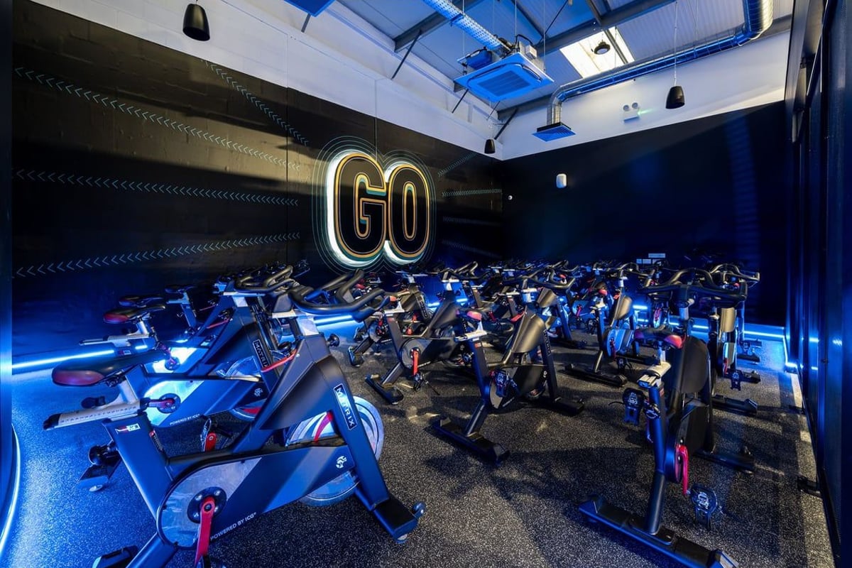 PureGym Newtownabbey: Rows of stationary cycles in dimly lit studio with neon 'GO' sign and blue lighting.