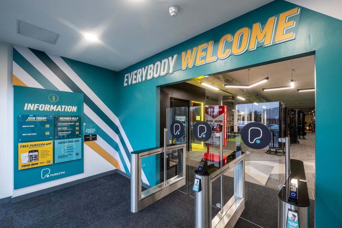 PureGym Derby Kingsway Retail Park: Entrance area with turnstiles, information signage, and "Everybody Welcome" wall mural.