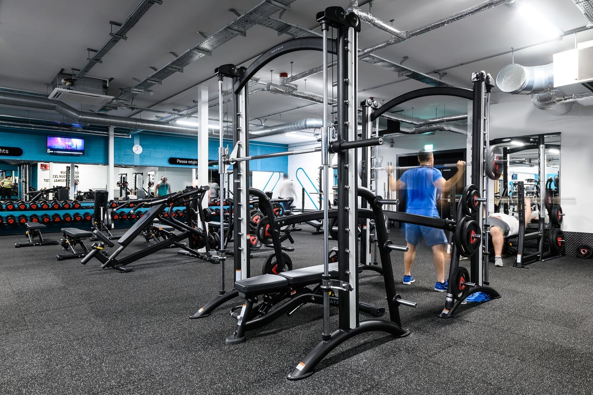 PureGym Cardiff Bay: Power racks, benches, barbells, weight plates, and rubberized flooring in a gym setting.