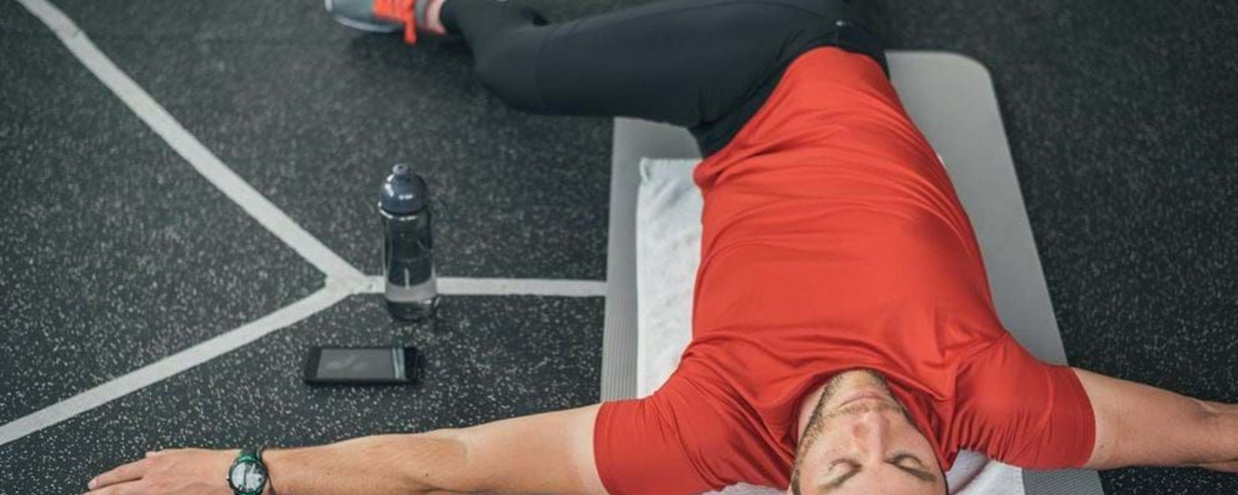 Man in red shirt stretching on mat with water bottle and phone nearby