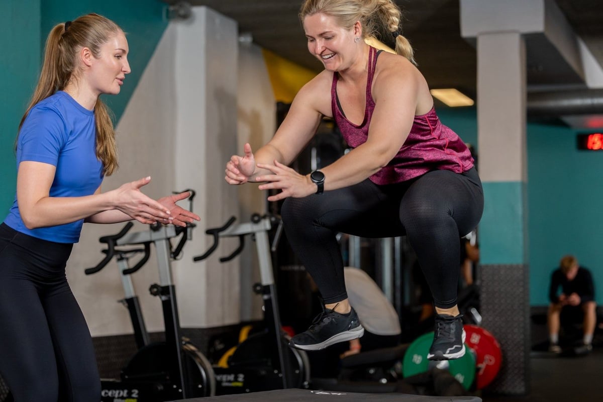 Woman in purple top performing box jump exercise whilst trainer in blue top observes
