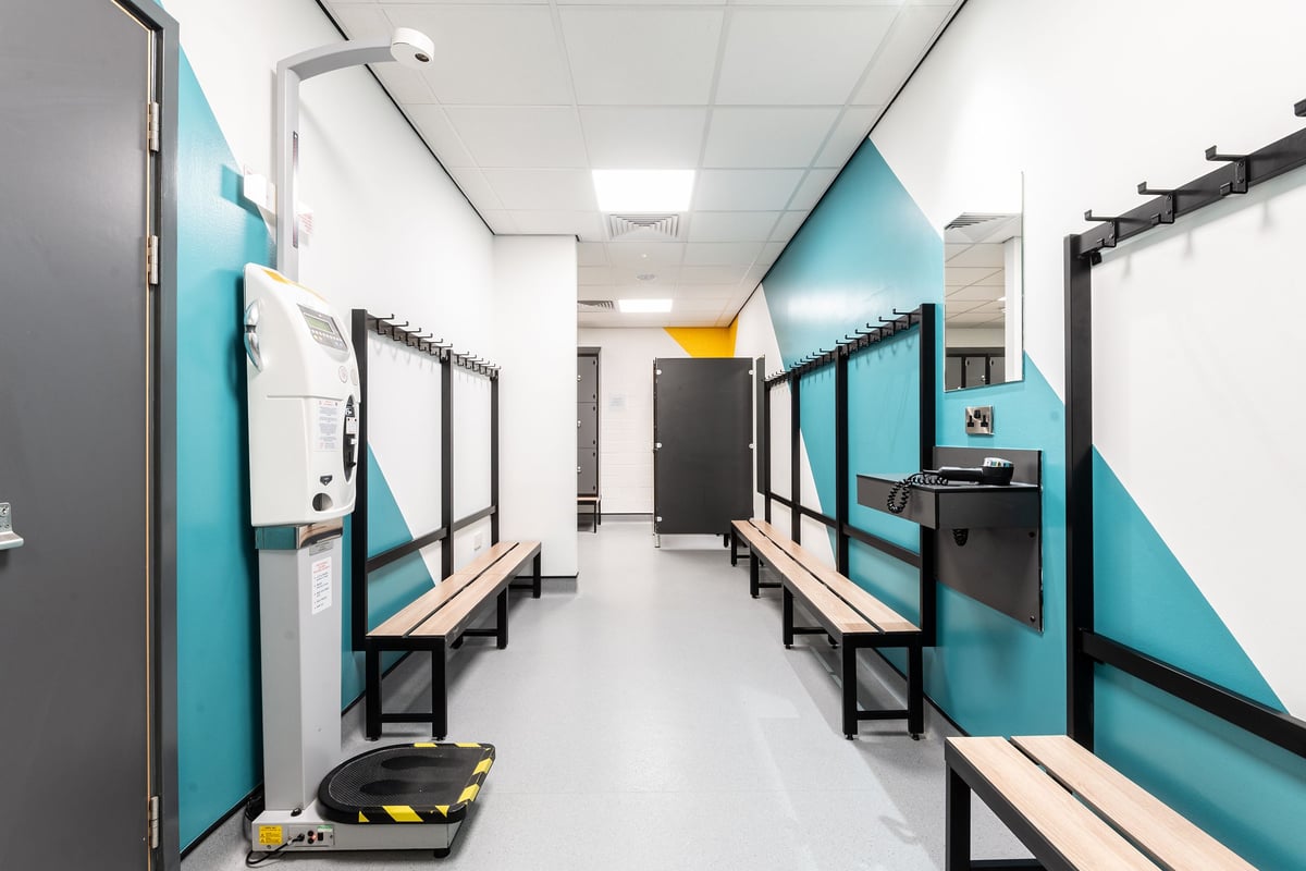 PureGym Banbury Cross Retail Park: Indoor changing rooms with benches, lockers, and cubicles in teal and grey.
