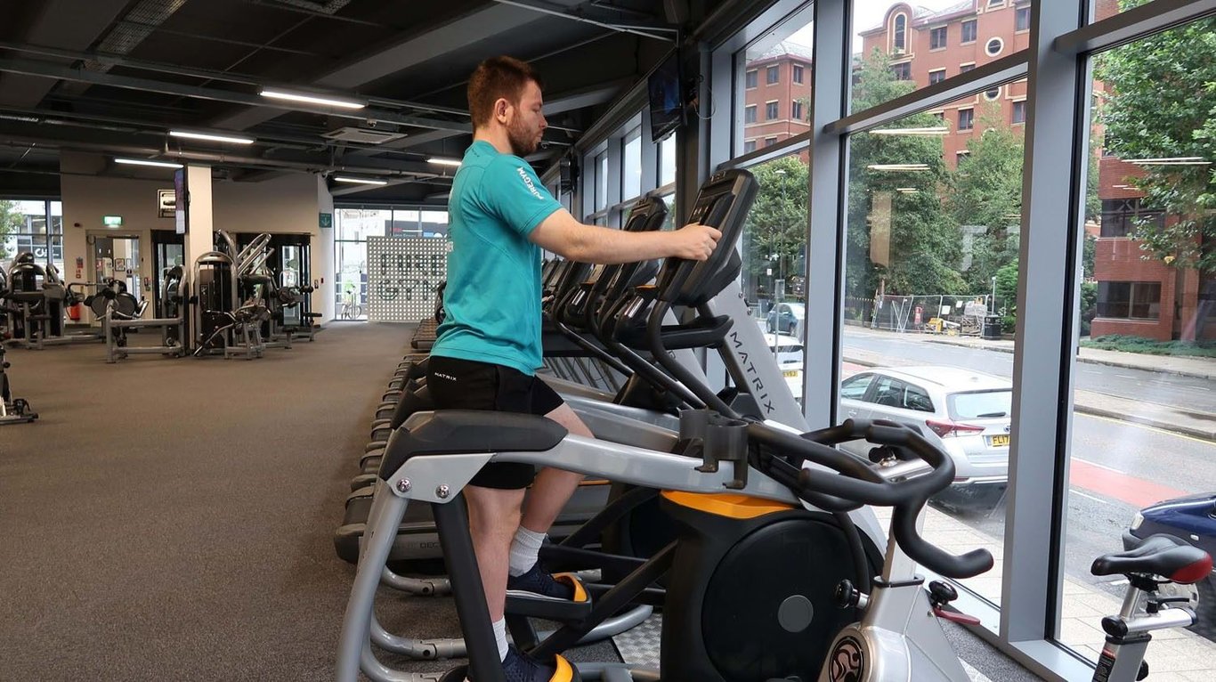 Man exercising on a stepper machine in a gym with large windows overlooking the street