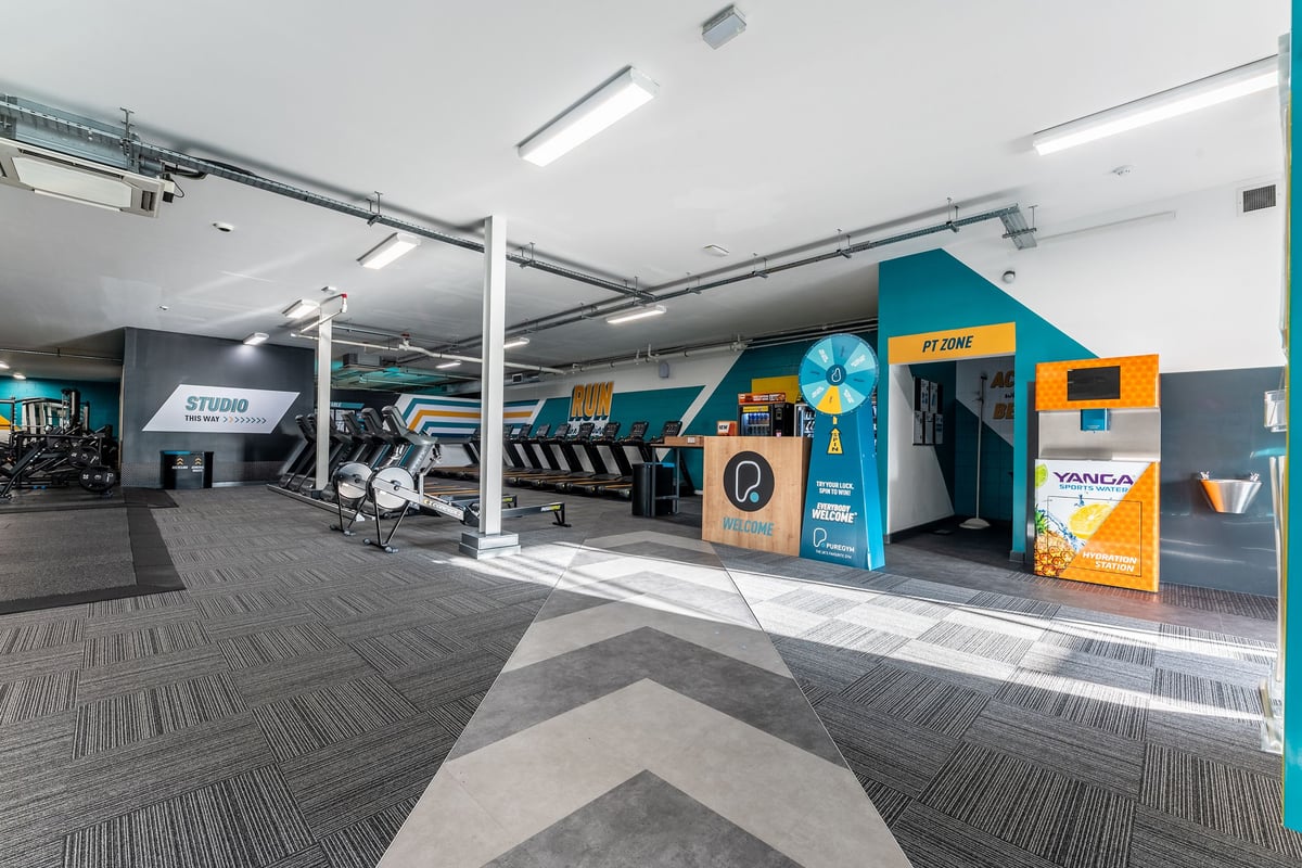 PureGym Ashton Under Lyne: Spin bikes, weightlifting area, stretch mats, and self-service kiosks in bright gym interior.