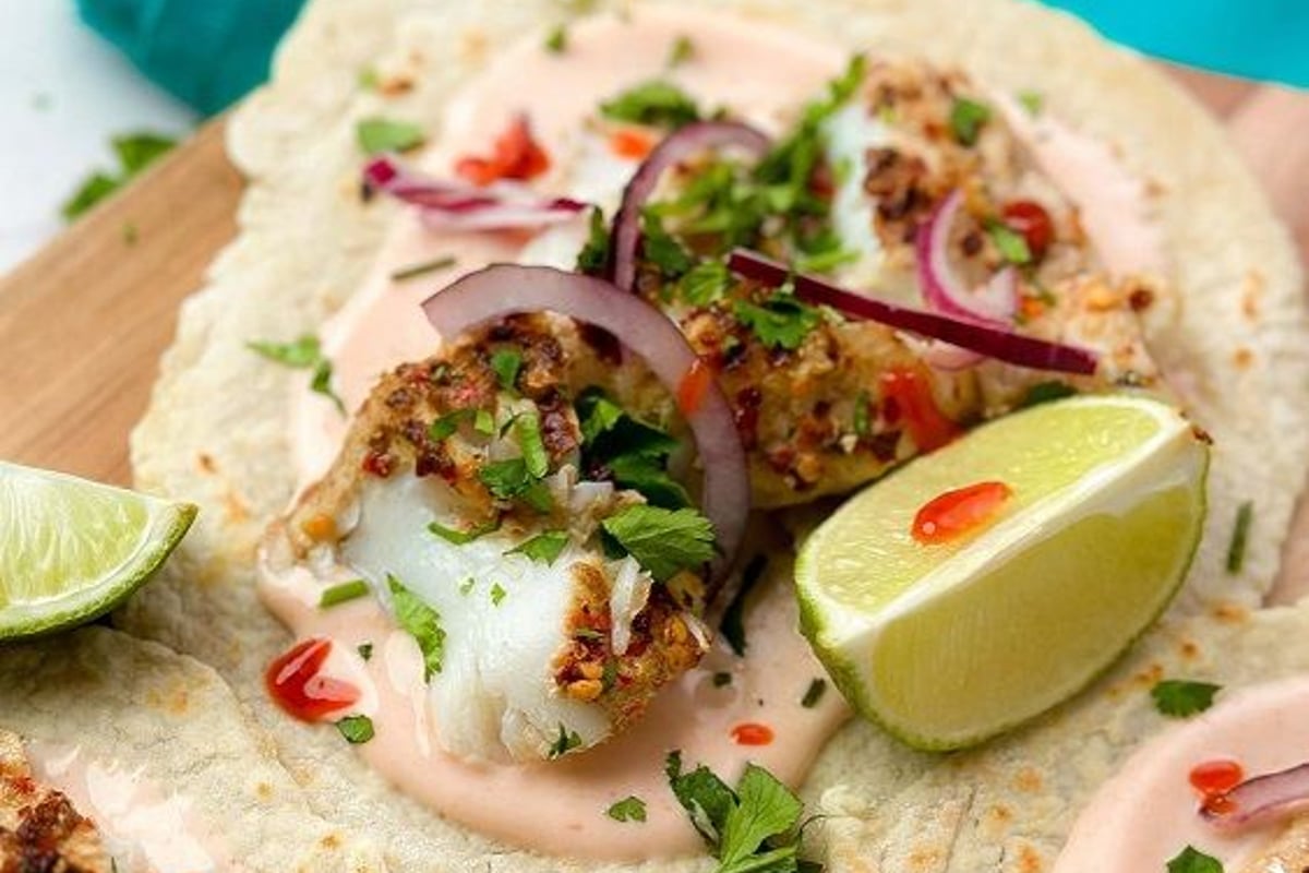 Fish tacos with sauce, red onion, herbs, and lime wedges on a wooden board