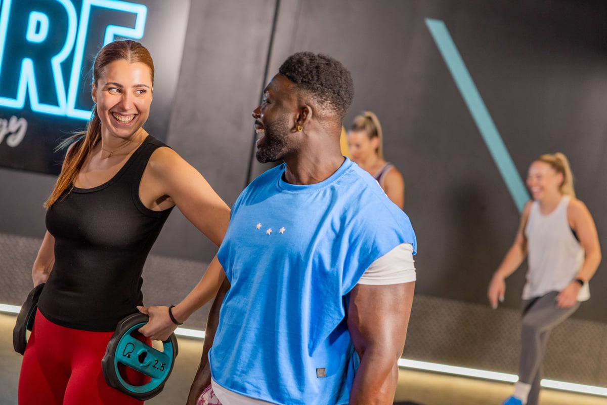 Two people smiling and holding weights in a gym with two others exercising behind them