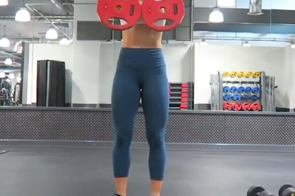 Woman lifting two red weight plates in a gym with dumbbells on the floor nearby