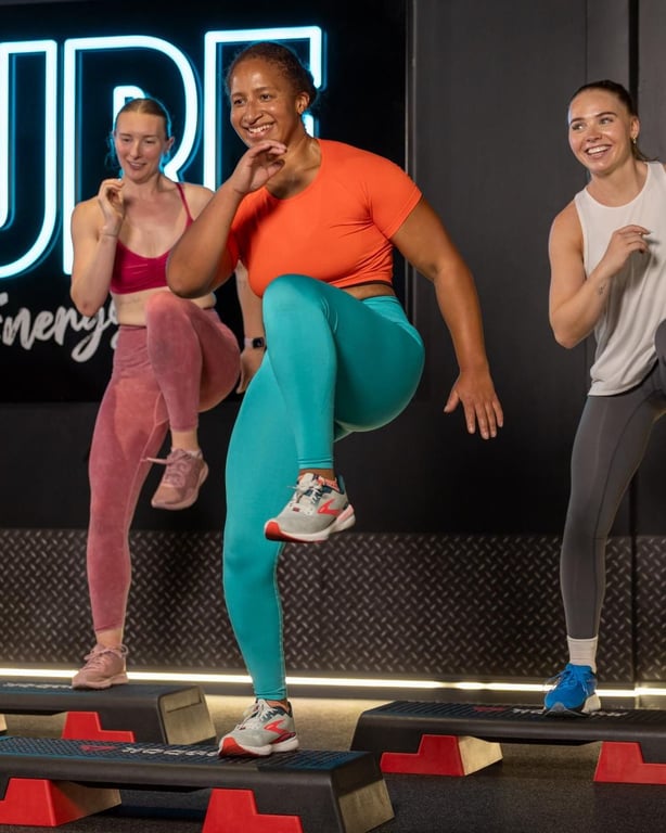 Drei lächelnde Frauen in farbenfroher Sportbekleidung trainieren auf Plattformen mit leuchtendem PureGym-Schriftzug im Hintergrund