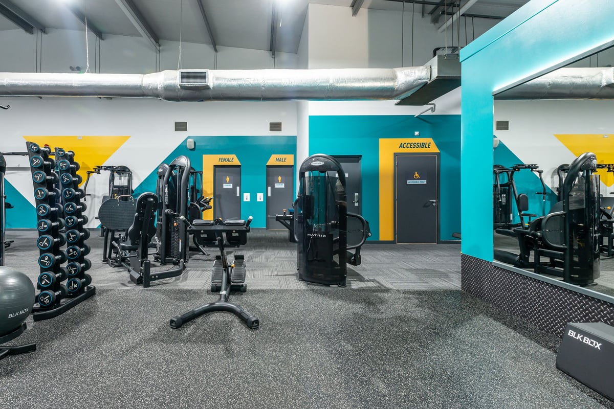 PureGym Wrexham: Strength training area with weight machines, free weights racks, benches, and rubberized flooring.