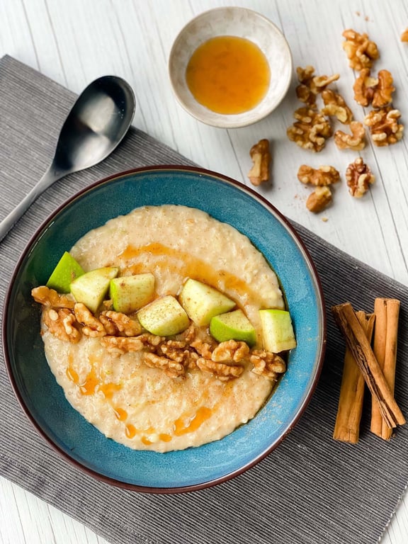 Bowl of porridge topped with walnuts, apple chunks, honey, and cinnamon sticks nearby