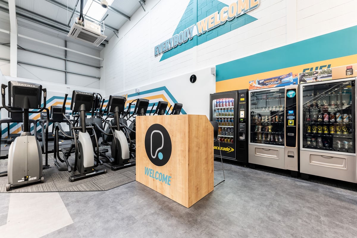 PureGym Blyth: Rows of exercise bikes, vending machines, welcome sign amidst turquoise branding.