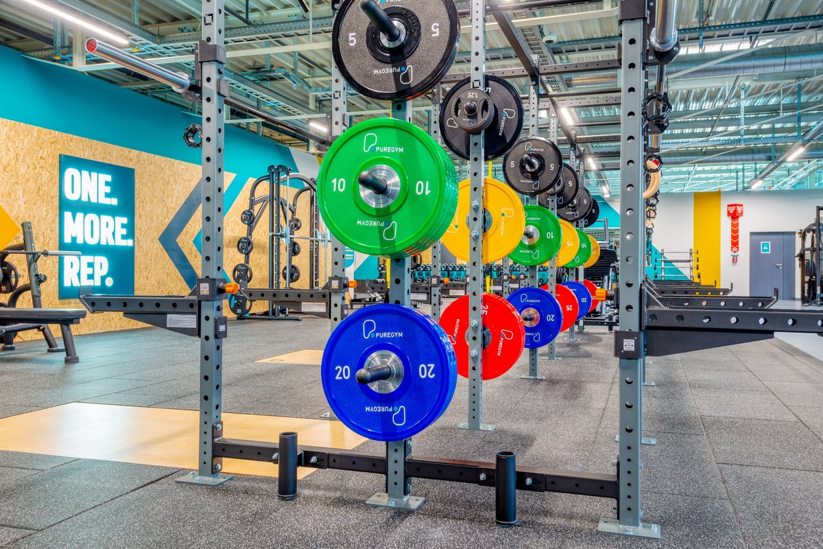 PureGym Martigny: Kniebeugen, mehrfarbige Hantelscheiben, die die Regale auskleiden, und gummierter Bodenbelag.