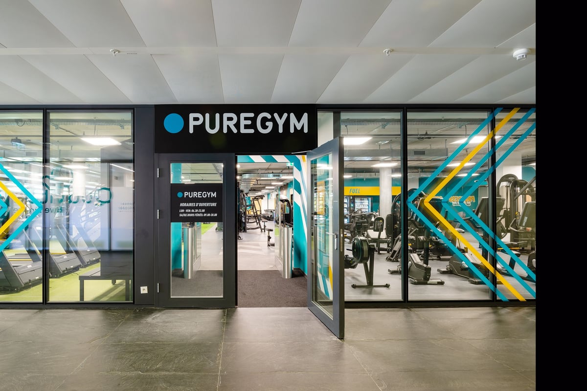 PureGym Genf Le Bouchet: PureGym entrance with glass doors, exercise equipment visible inside, and colourful wall graphics.