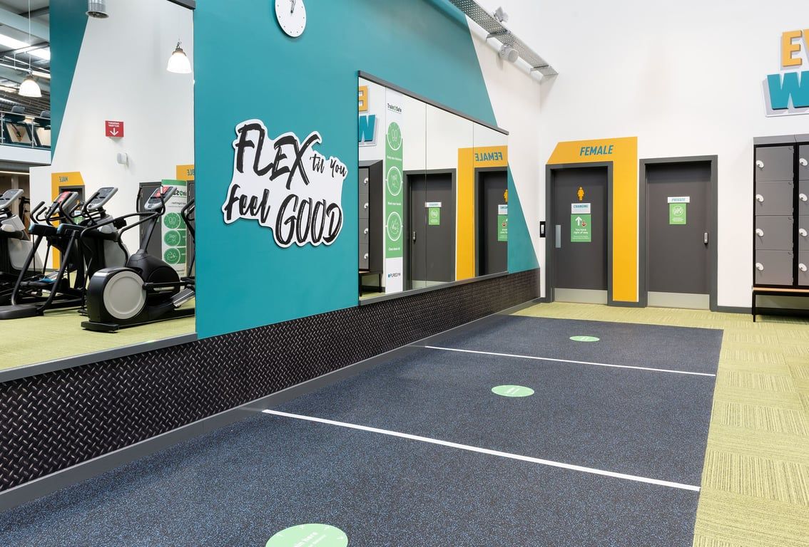 PureGym Colchester Retail Park: Rows of exercise bikes, gym turf flooring, strength equipment, and colorful wall graphic.